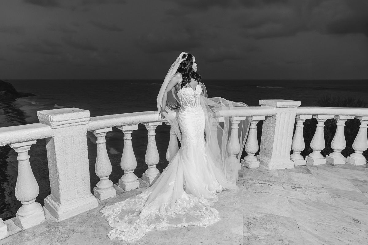 Bride in elegant mermaid wedding gown with veil poses on white balustrade terrace overlooking ocean in black and white photo.