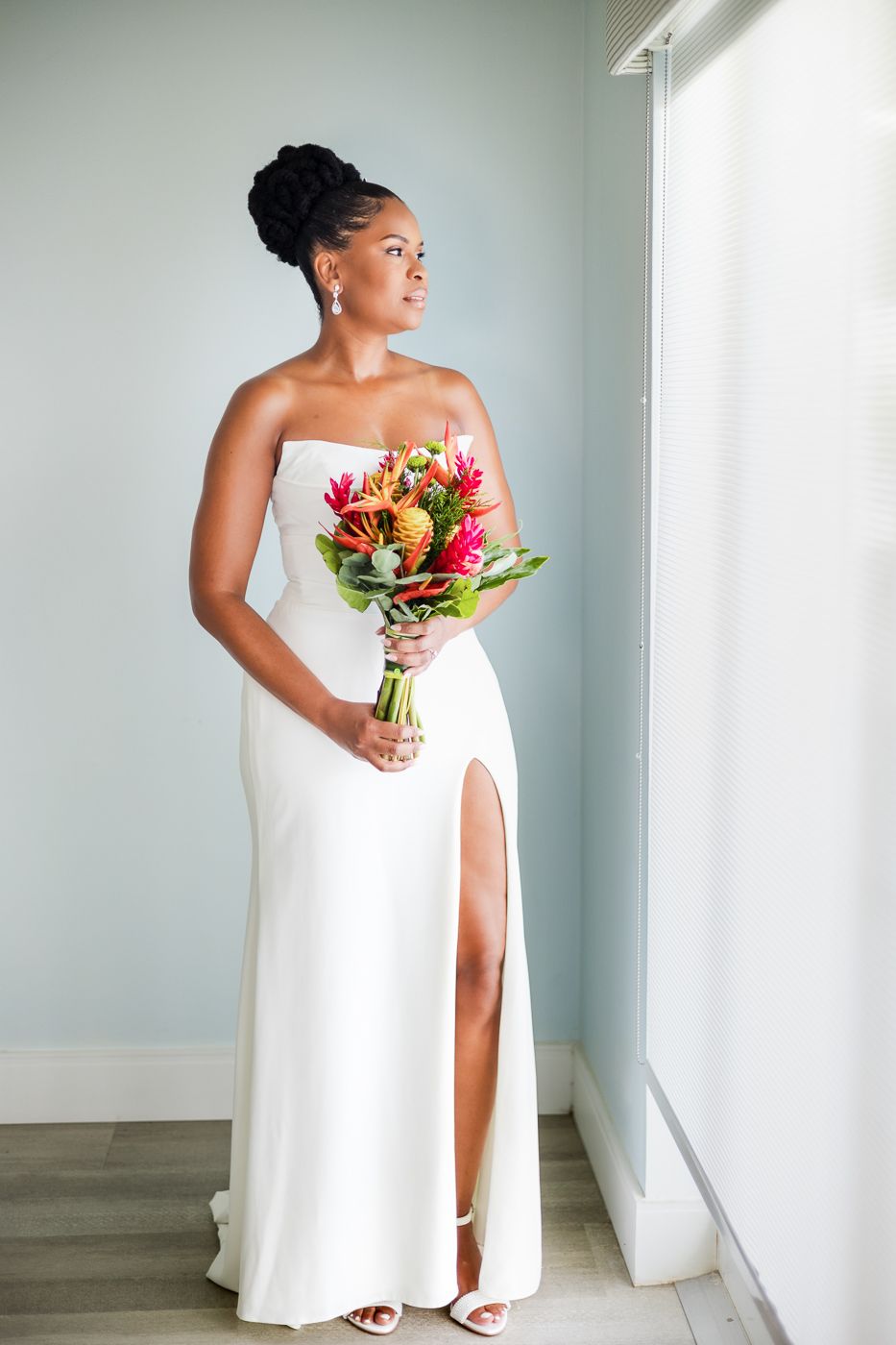 Elegant bride in white strapless gown holding tropical bouquet, standing by window in soft natural light.