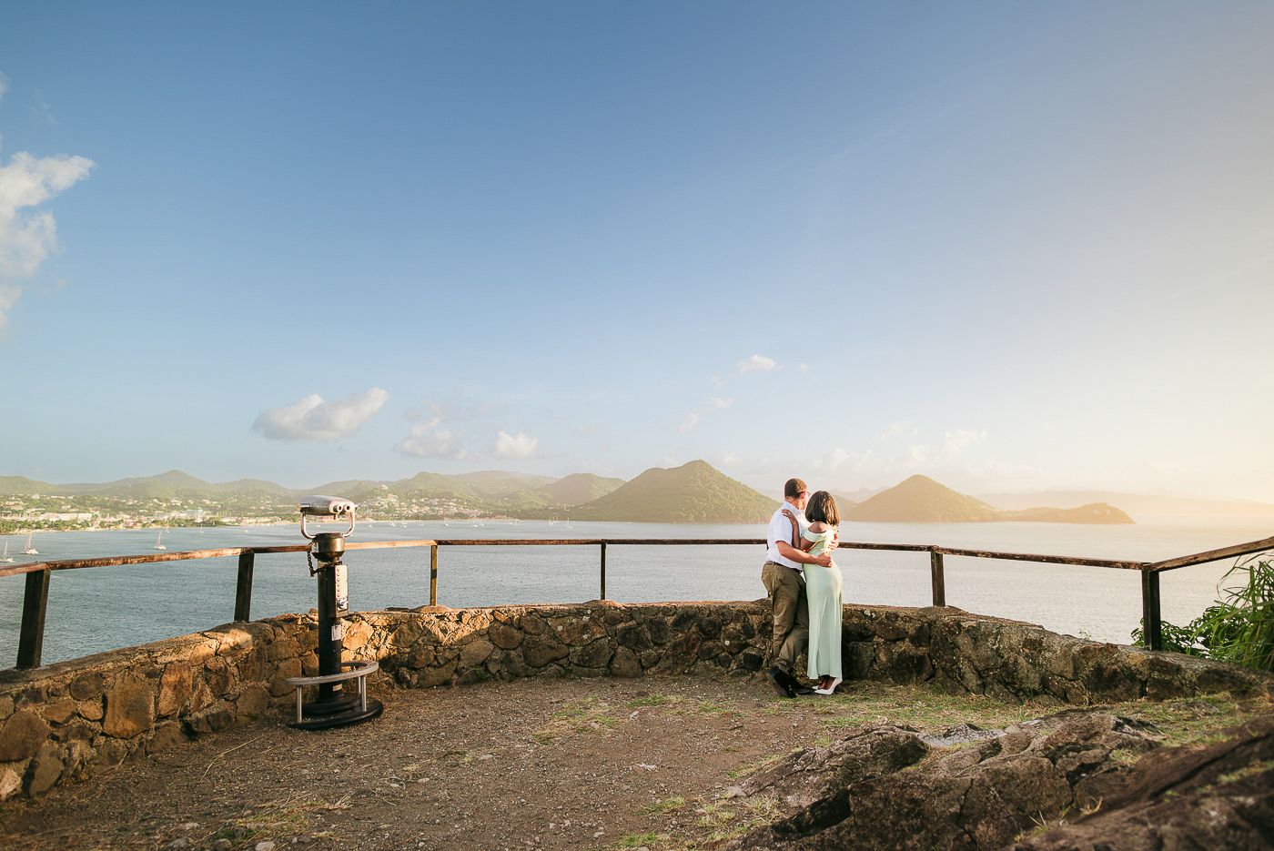 Couple embracing at a scenic overlook with ocean and mountain views under a golden sunset sky.