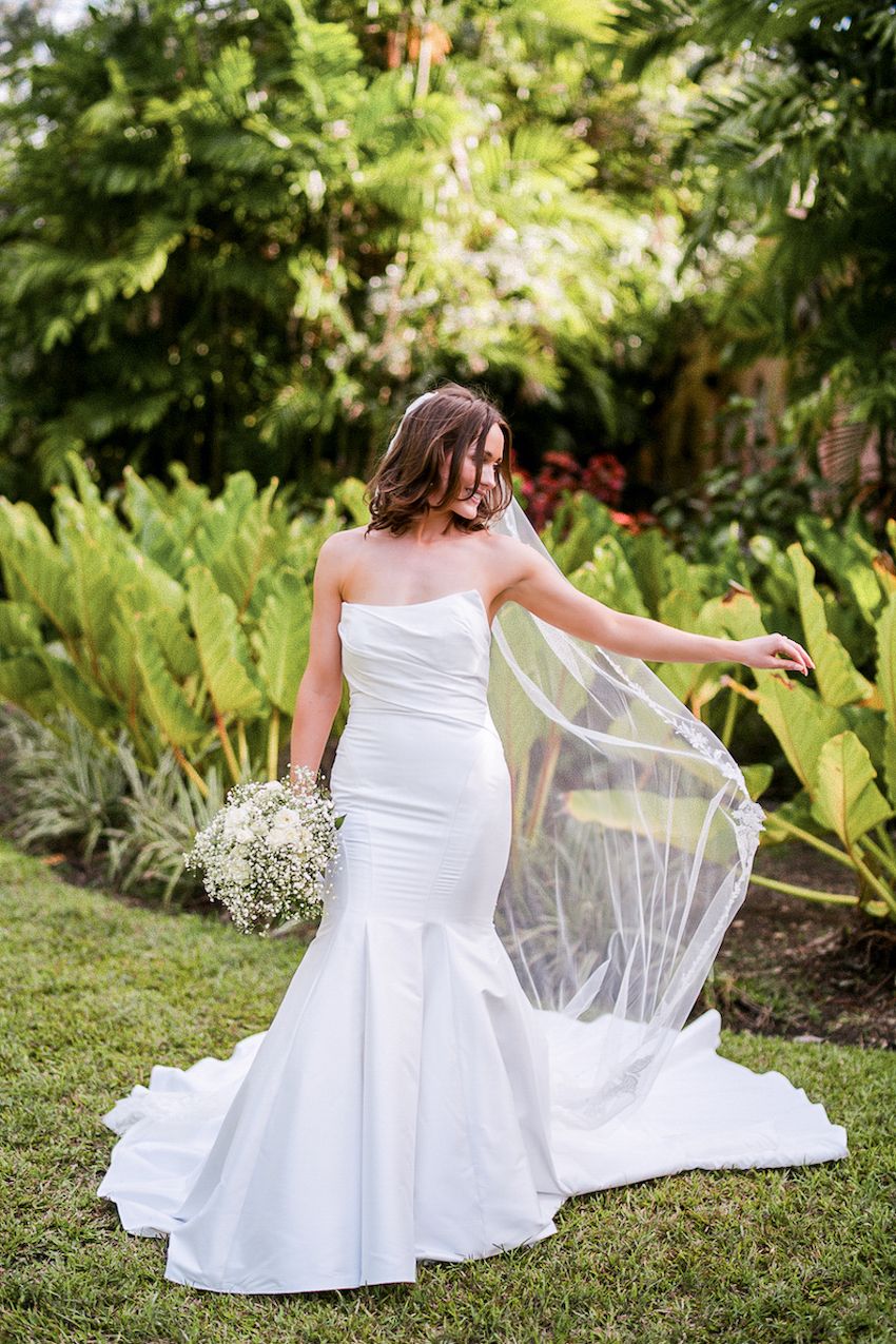 Bride in strapless mermaid wedding gown holding bouquet, veil flowing in tropical garden setting.