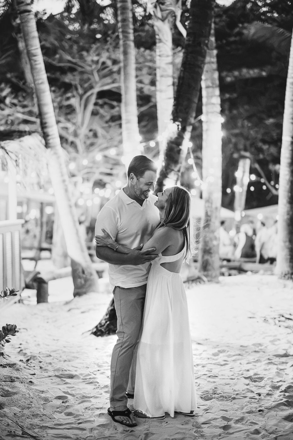Black and white photo of a couple embracing and smiling on a sandy beach surrounded by palm trees and string lights.