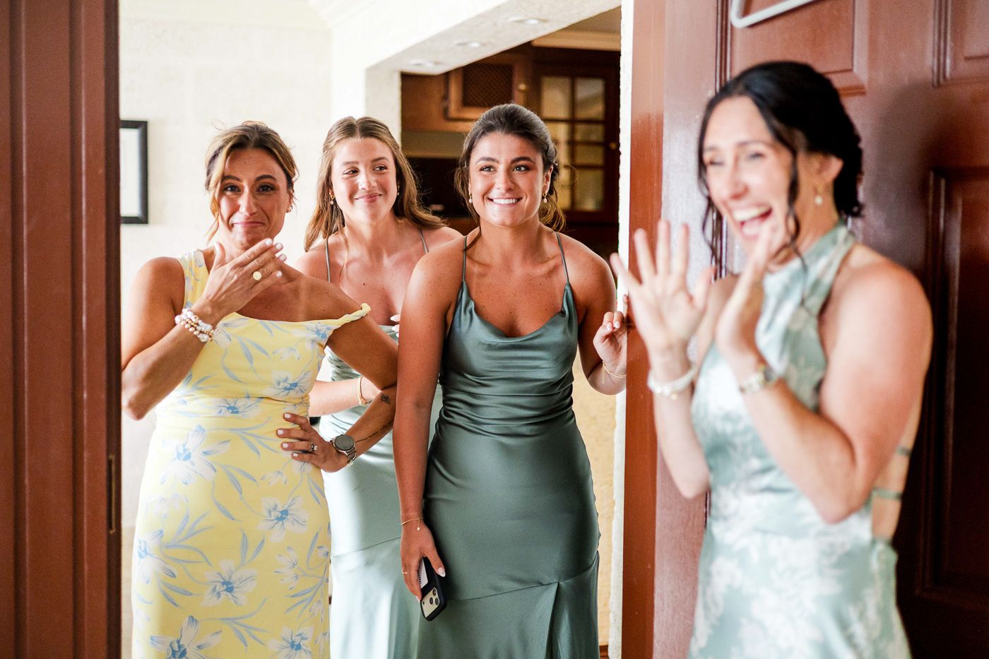 Four women in elegant dresses smile and show off rings, celebrating a wedding day together.