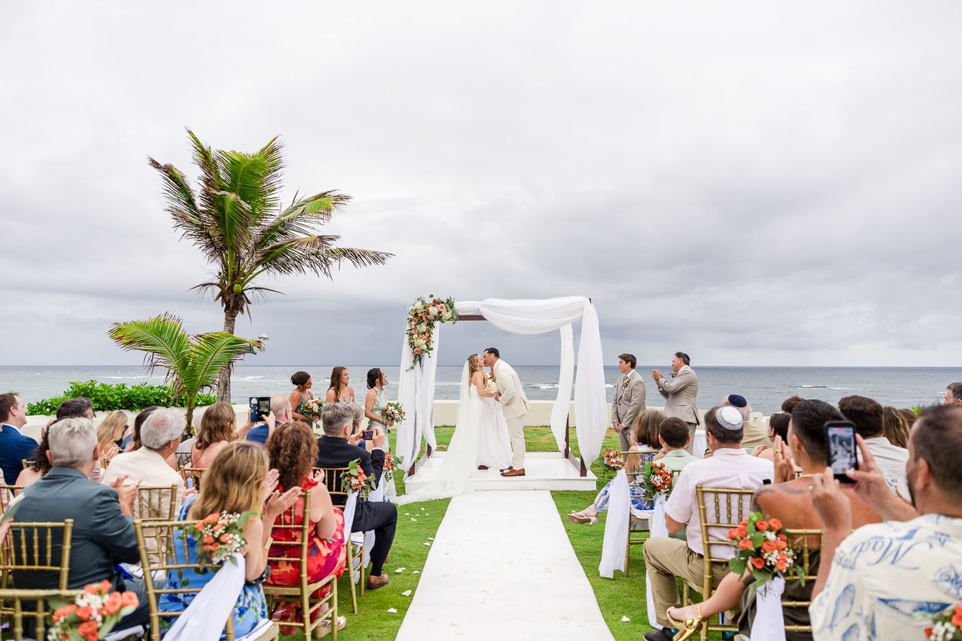 Outdoor beach wedding ceremony with couple kissing at floral arch, guests seated on gold chairs, palm tree and ocean backdrop.