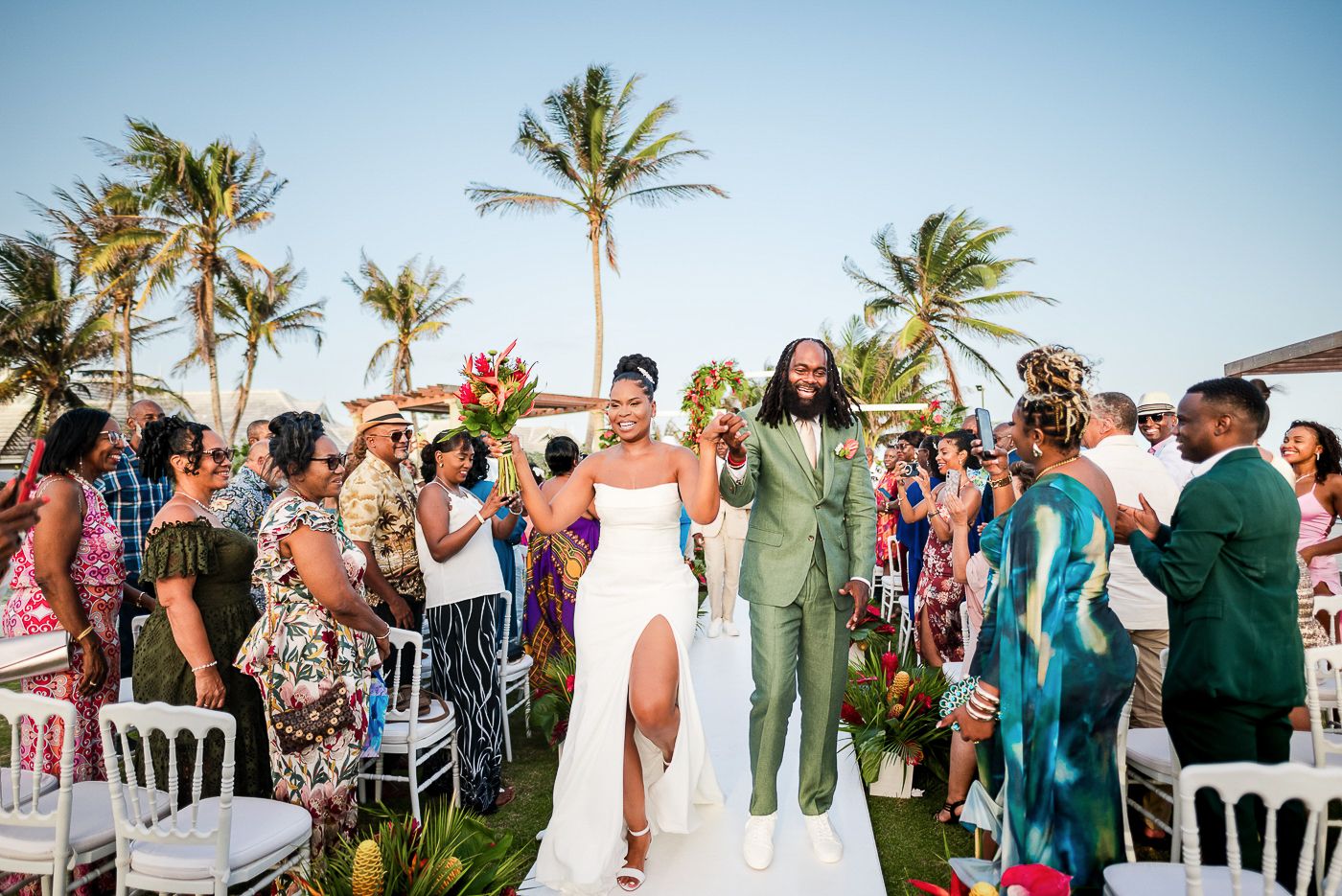 Tropical beach wedding ceremony with palm trees and guests in colorful attire.