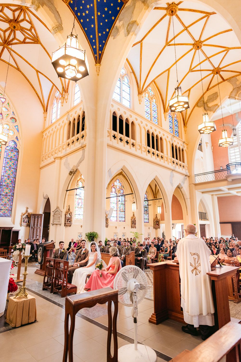 Panoramic wedding ceremony in a grand cathedral with high vaulted ceilings, stained glass windows and ornate chandeliers.