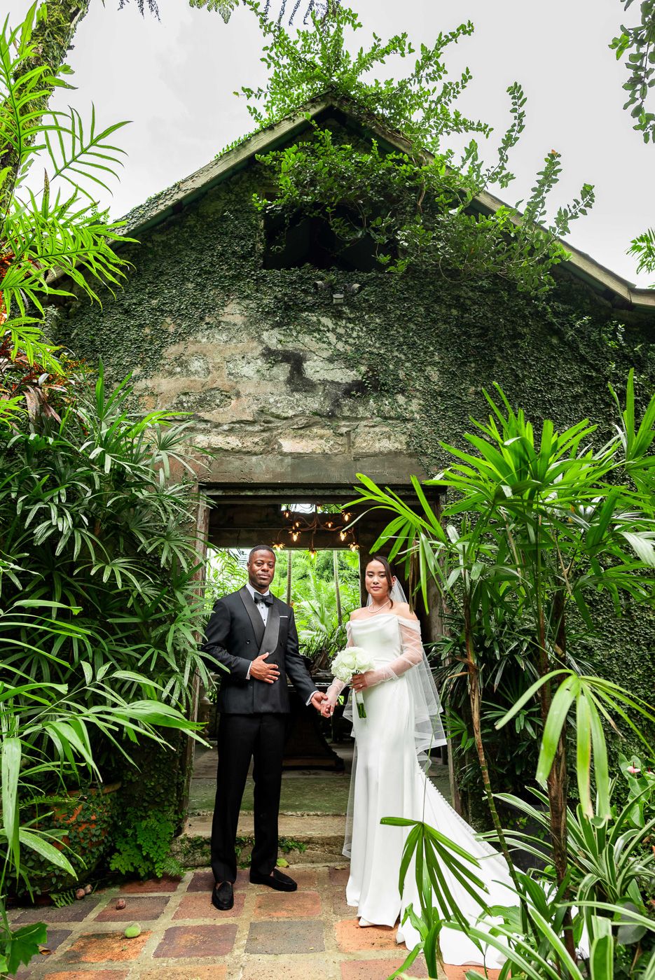 Wedding couple poses in doorway of vine-covered stone building surrounded by tropical plants.