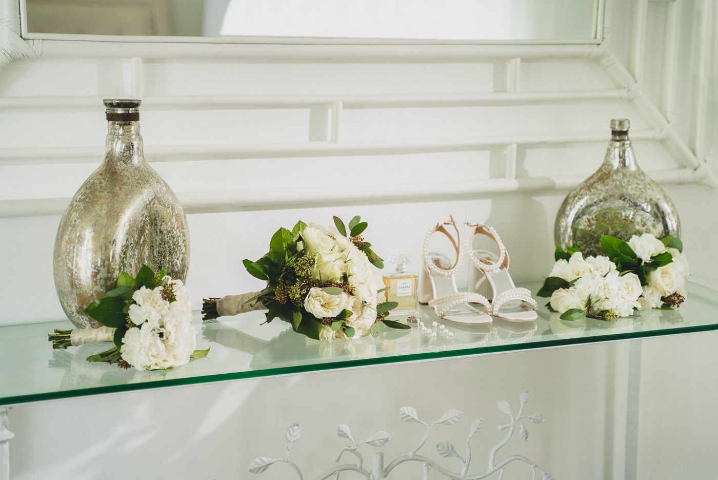 Glass vases and wedding accessories arranged on a glass shelf with white roses and decorative elements.