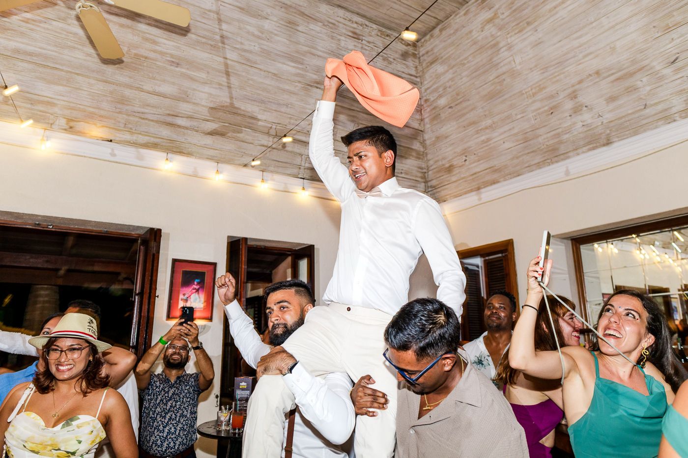 Energetic party scene with guests dancing and jumping under wooden beams and string lights.