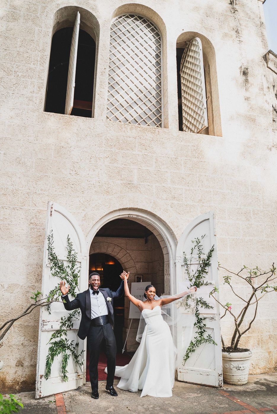 Wedding couple celebrating at church entrance with white stone walls and decorative windows.