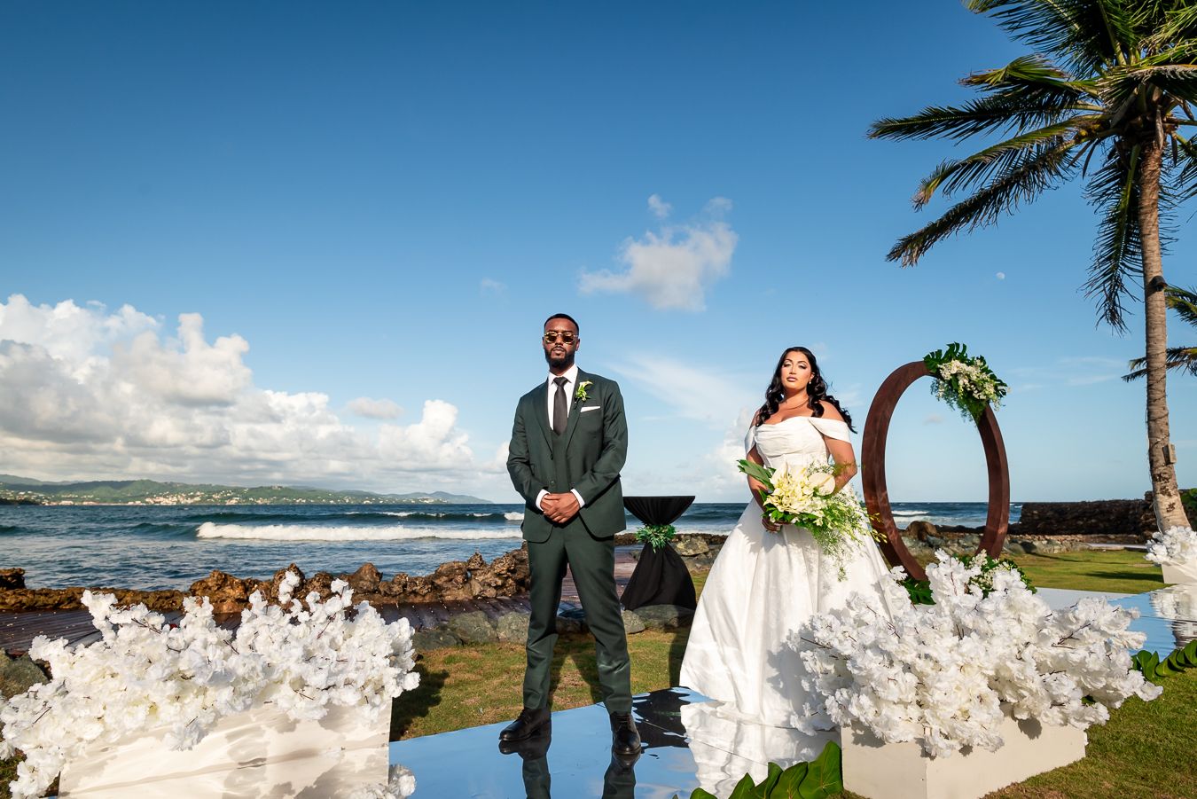 Wedding ceremony setup on tropical beachfront with palm trees and white chairs.