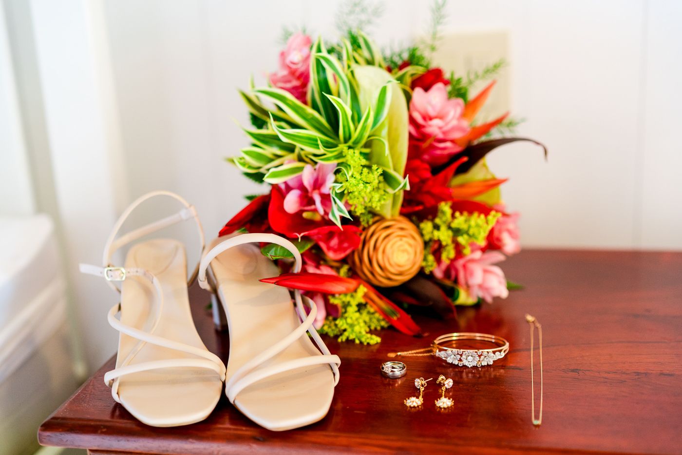 Bridal accessories including strappy sandals, wedding rings, and vibrant red and pink floral bouquet.