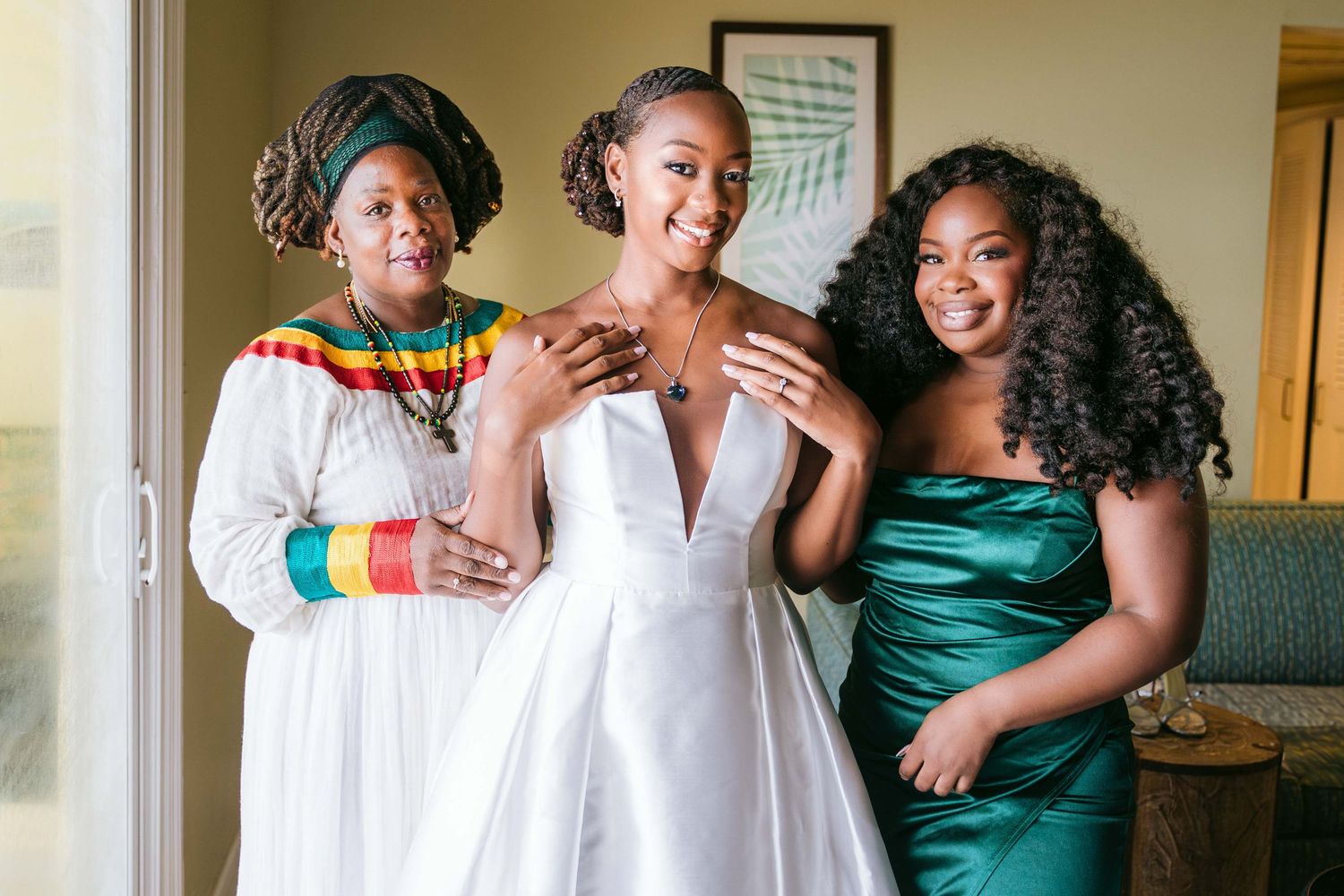 Three people share a joyful moment while getting ready, with one wearing a white wedding dress and others in colorful attire.
