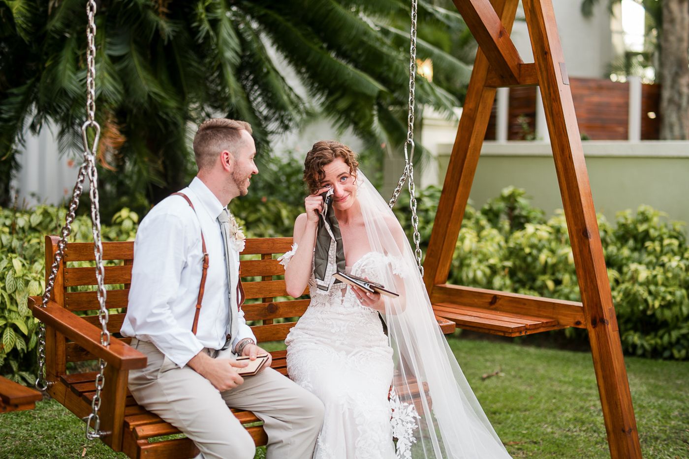 Newlyweds share intimate moments on wooden swing in garden setting.