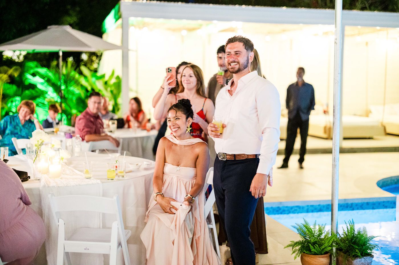 Wedding party and guests mingling during outdoor reception with pool and garden views.