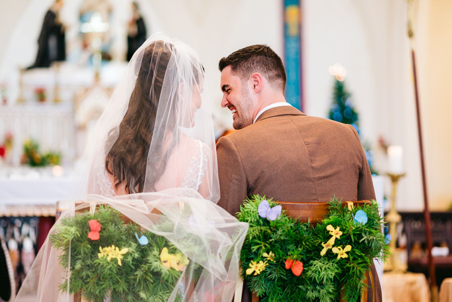 A couple dressed in wedding attire sits together at altar decorated with greenery and peach florals in a church.