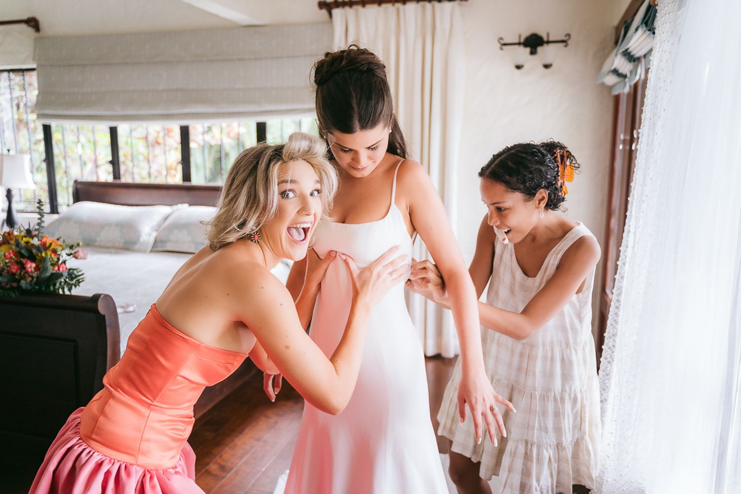 Bridesmaids help bride prepare for wedding day in bright room with white curtains and natural light.