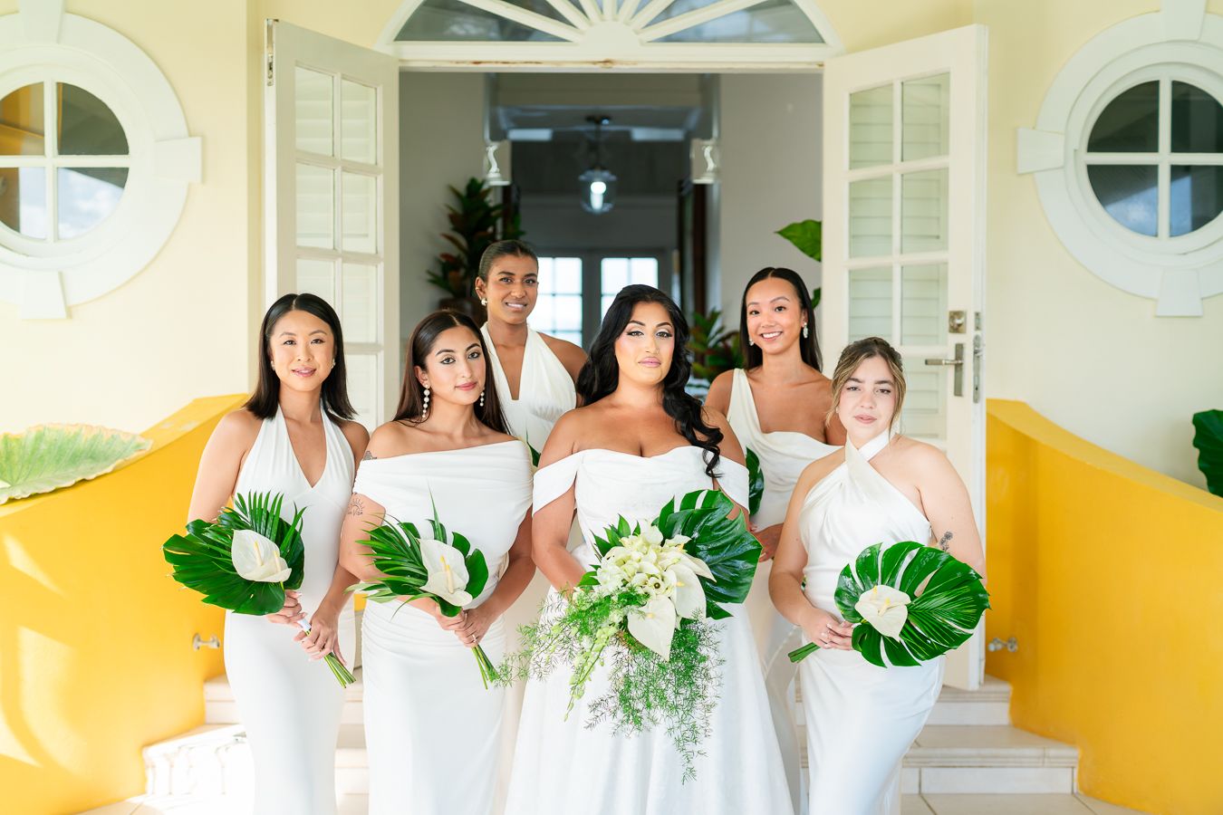 Bridesmaids in white dresses hold tropical leaf bouquets while posing in front of yellow walls.