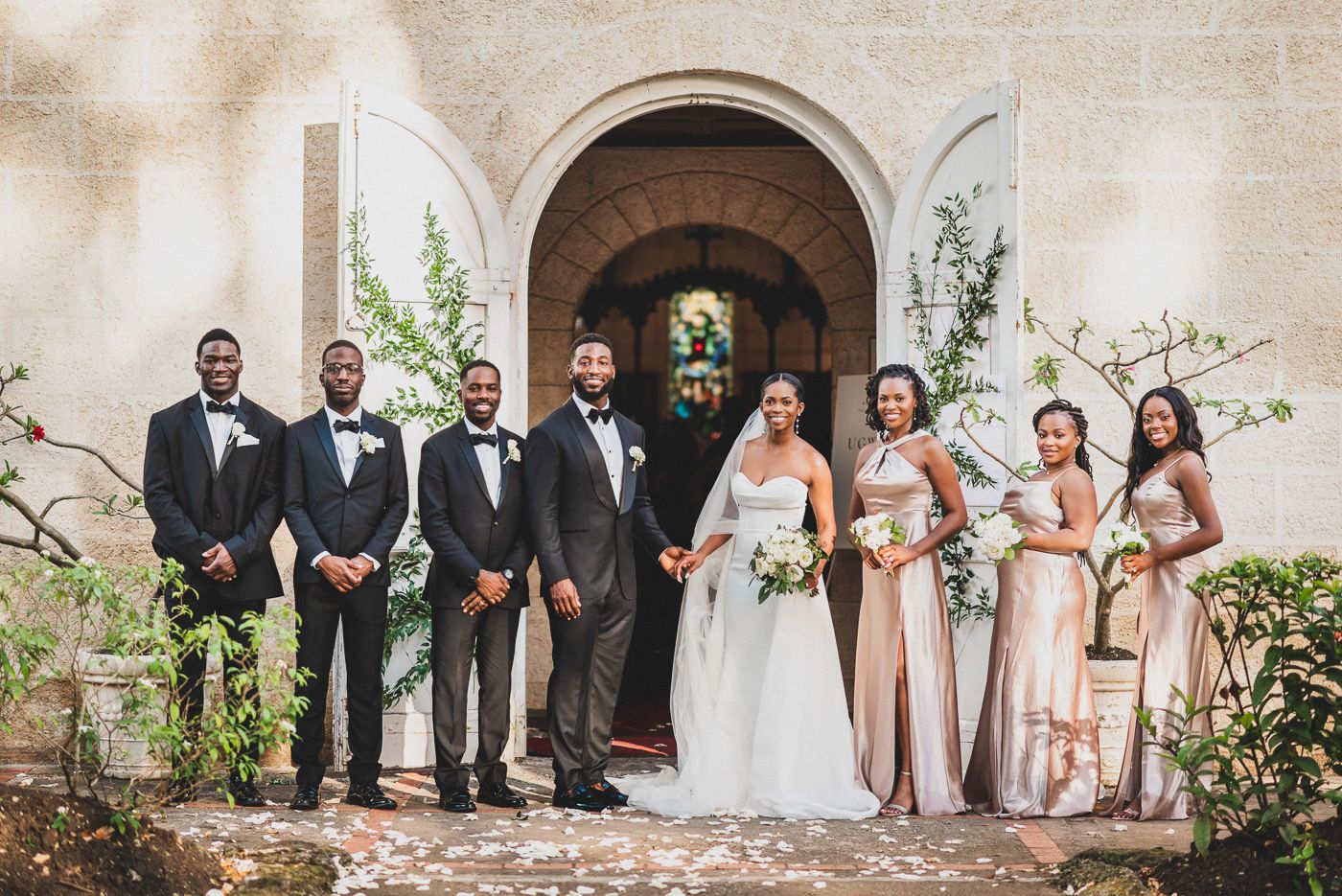 Wedding party lined up for formal portrait outside stone church entrance with floral decor.