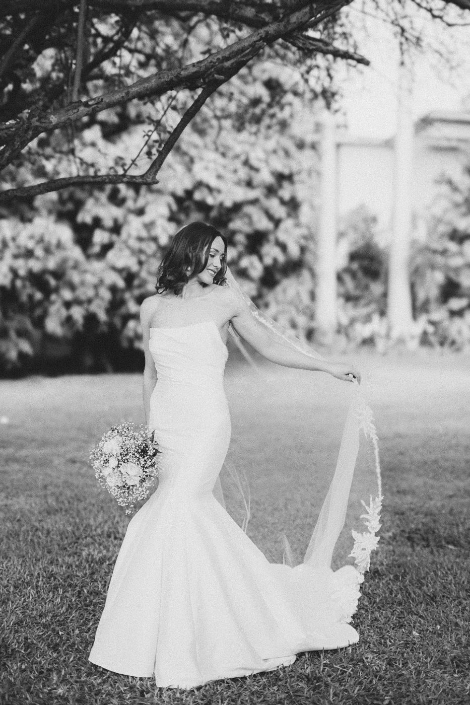 Black and white photo of bride twirling in classic white dress under garden trees.
