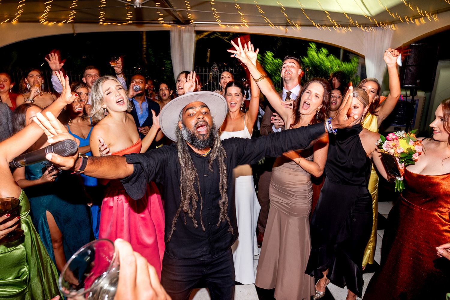 Wedding guests dancing and celebrating on a dance floor at an evening reception with their hands in the air.