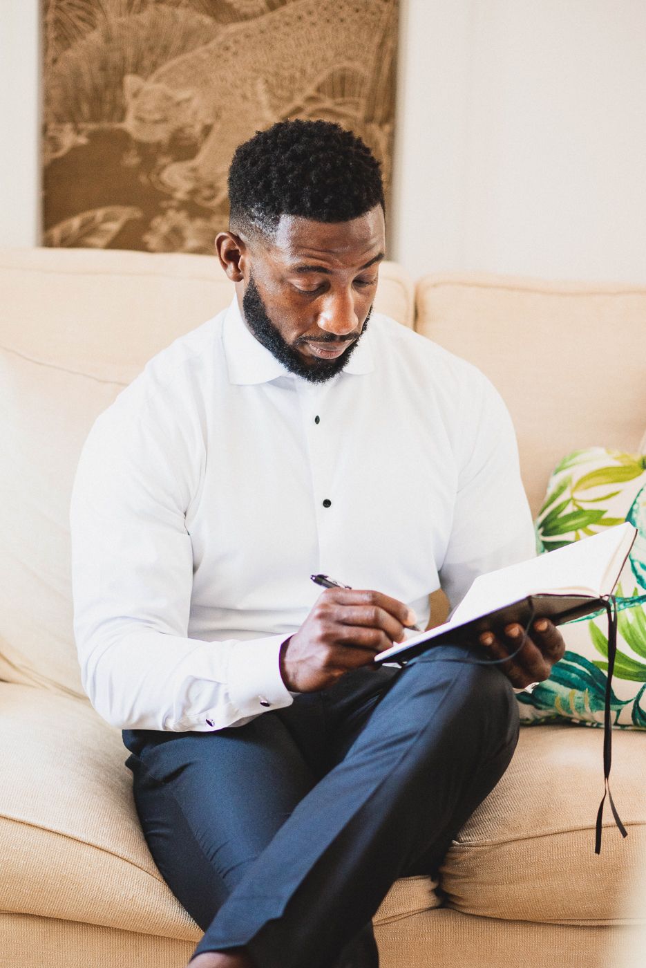 A person wearing a white button-down shirt sits on a beige couch writing in a journal.