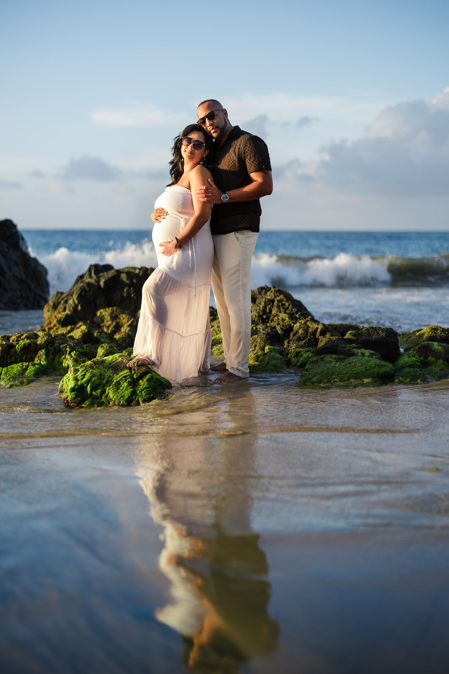 Pregnant couple embracing on a rocky beach at sunset, woman in white maternity gown, man in black shirt, ocean waves behind them.