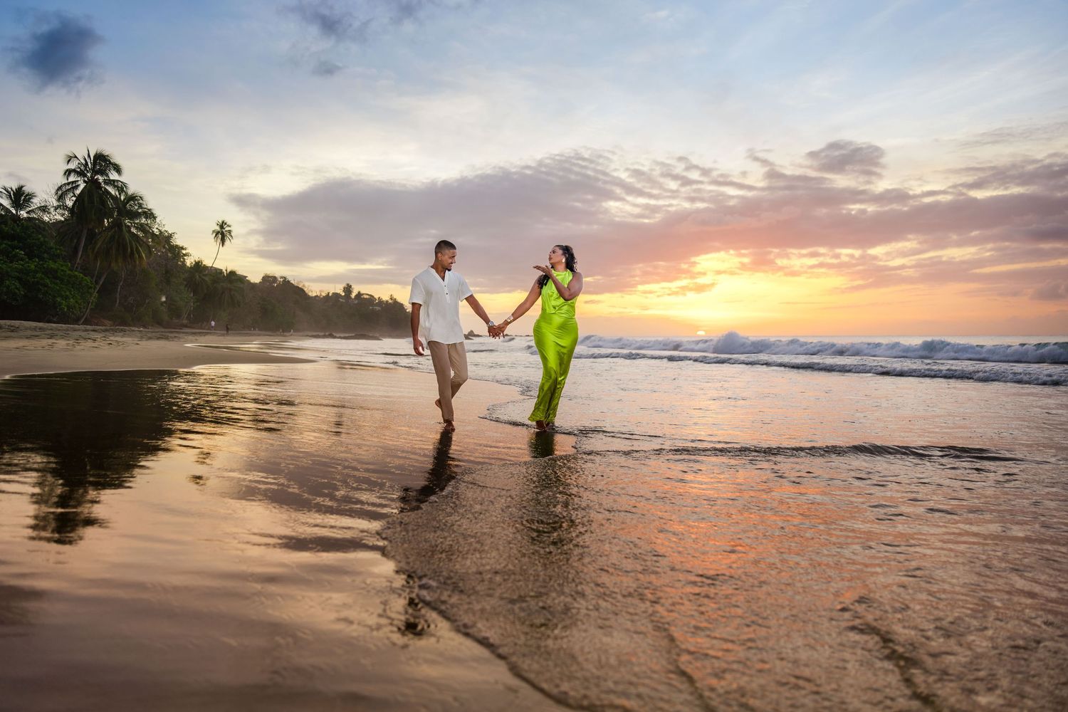 Couple holding hands walking on a tropical beach at sunset, woman in green sari, man in white shirt.