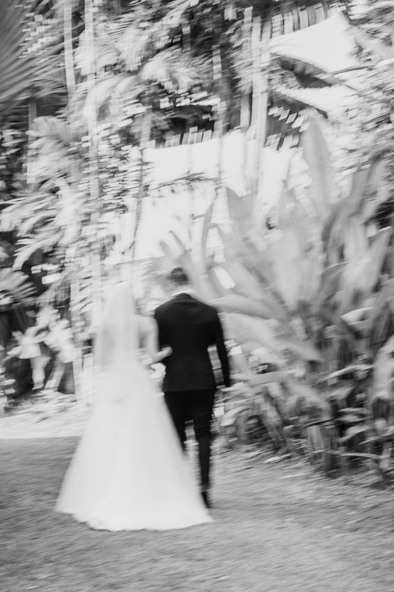 Black and white motion blur photograph of a couple in wedding attire walking through a garden setting.