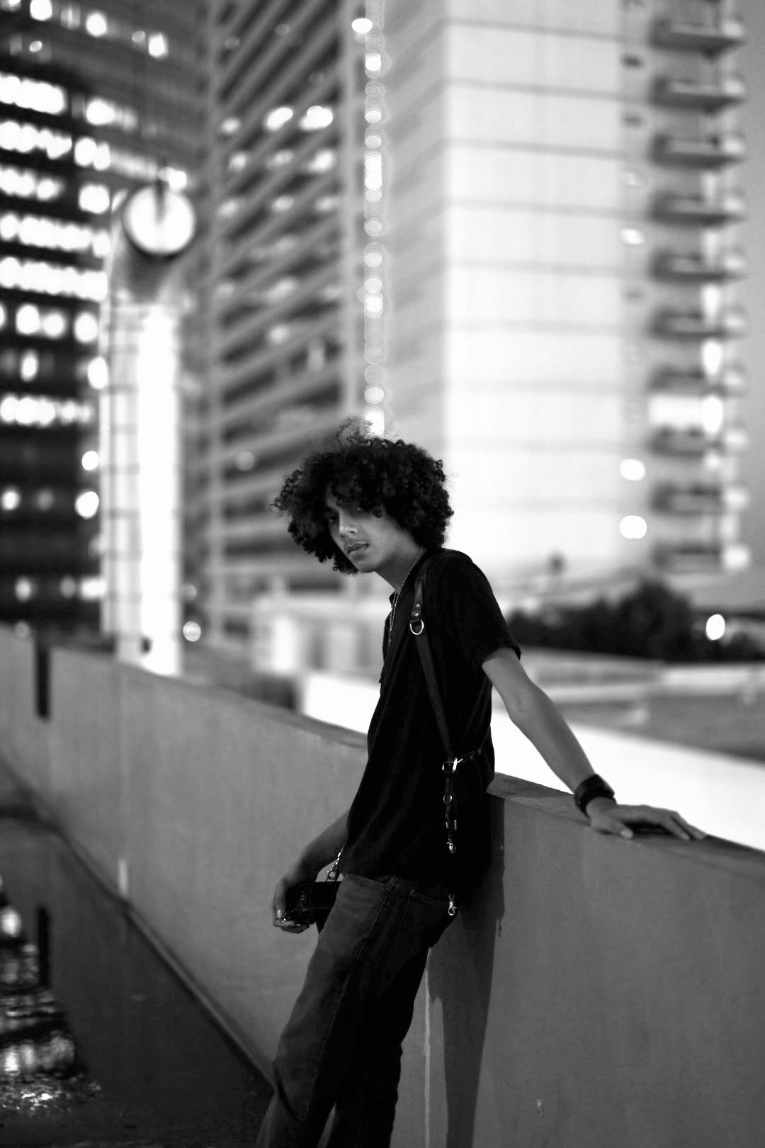 A figure in black clothing stands against a blurred cityscape background with bokeh lights in a monochrome composition.