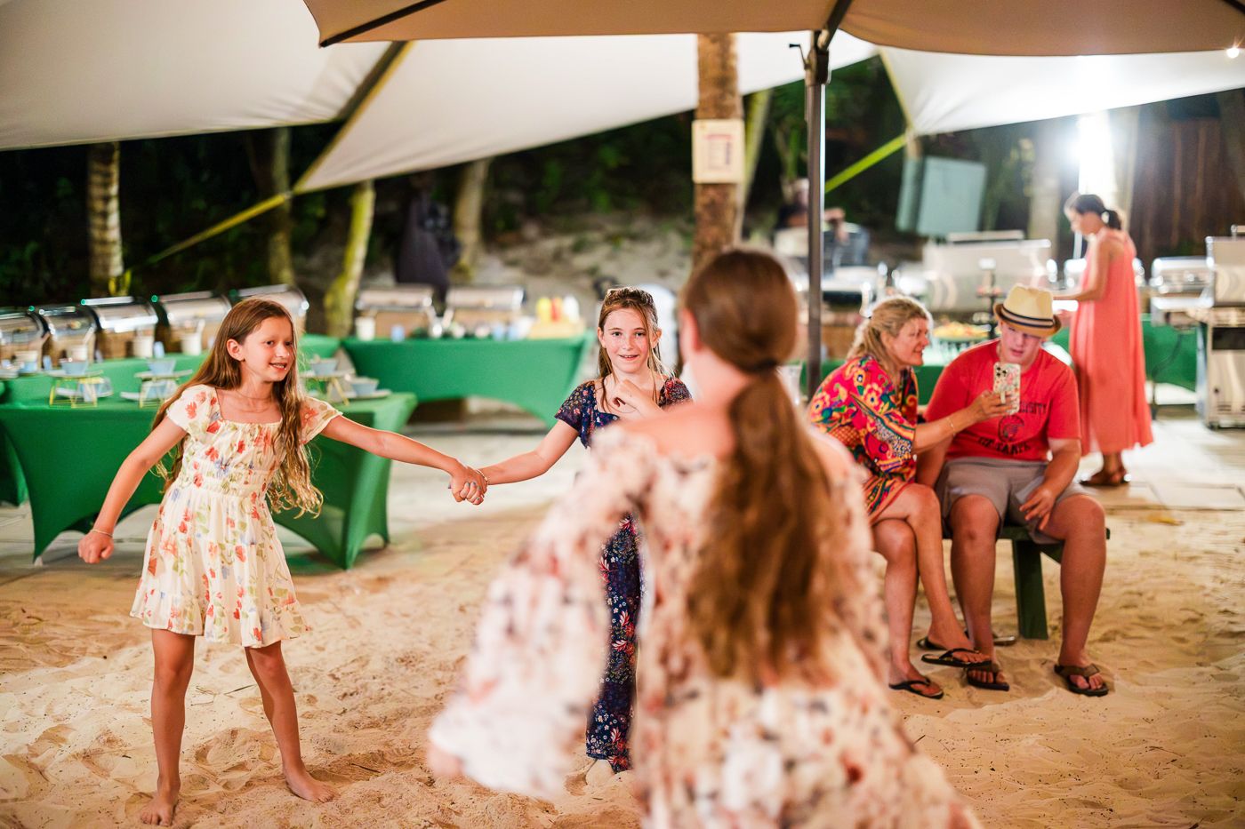 Children dancing joyfully at an outdoor beach party under a tent with guests seated nearby.