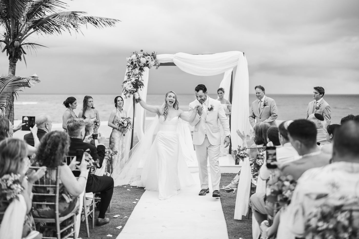 Joyful newlyweds celebrate at an outdoor beach wedding ceremony beneath a floral arch with guests cheering.