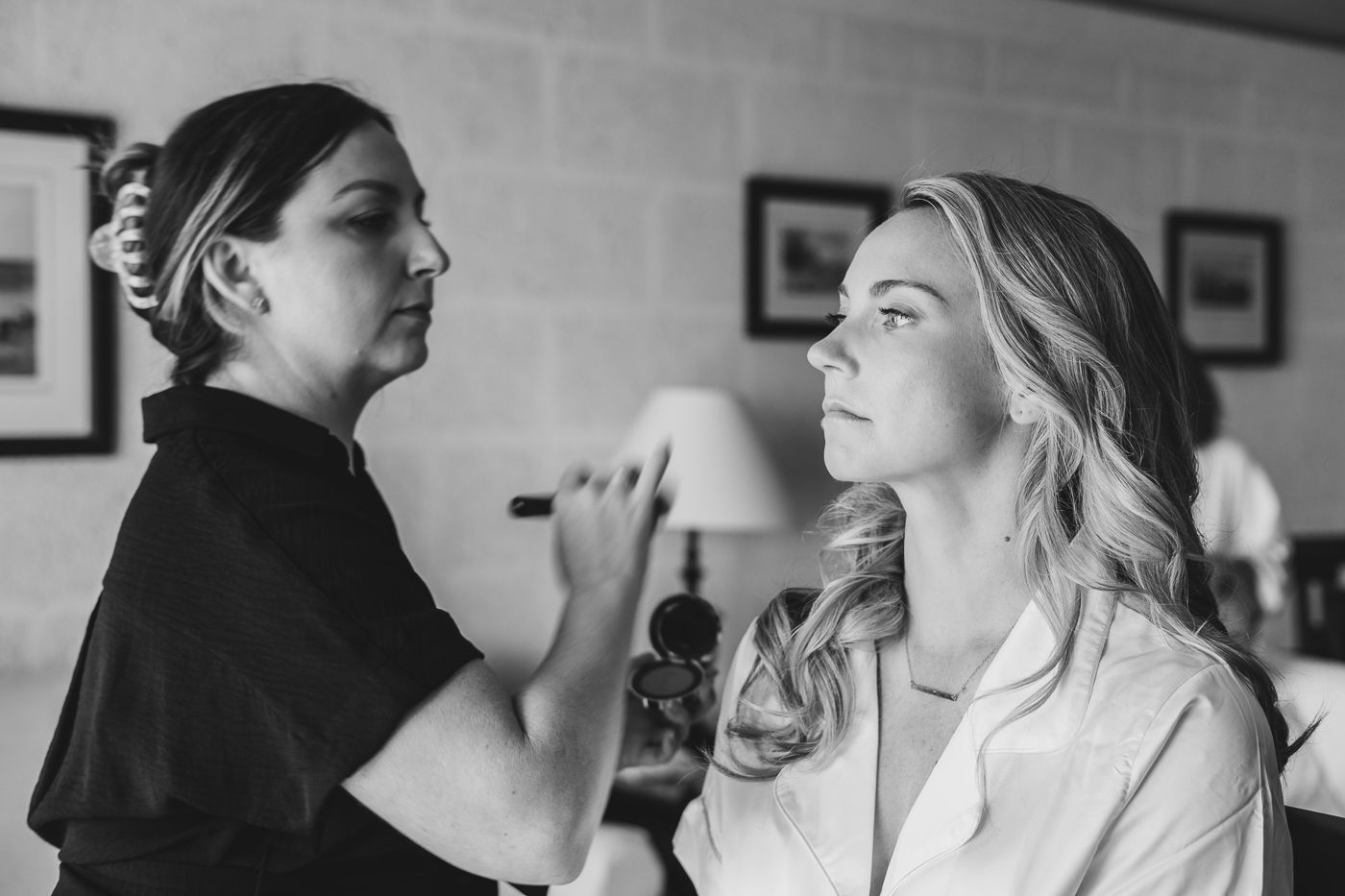 Black and white photo of a makeup artist applying makeup to a blonde bride during wedding preparations.