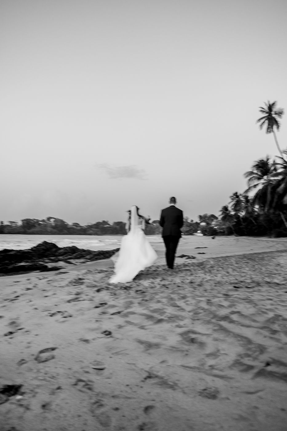 Black and white photo of a bride and groom walking hand in hand on a tropical beach at sunset.