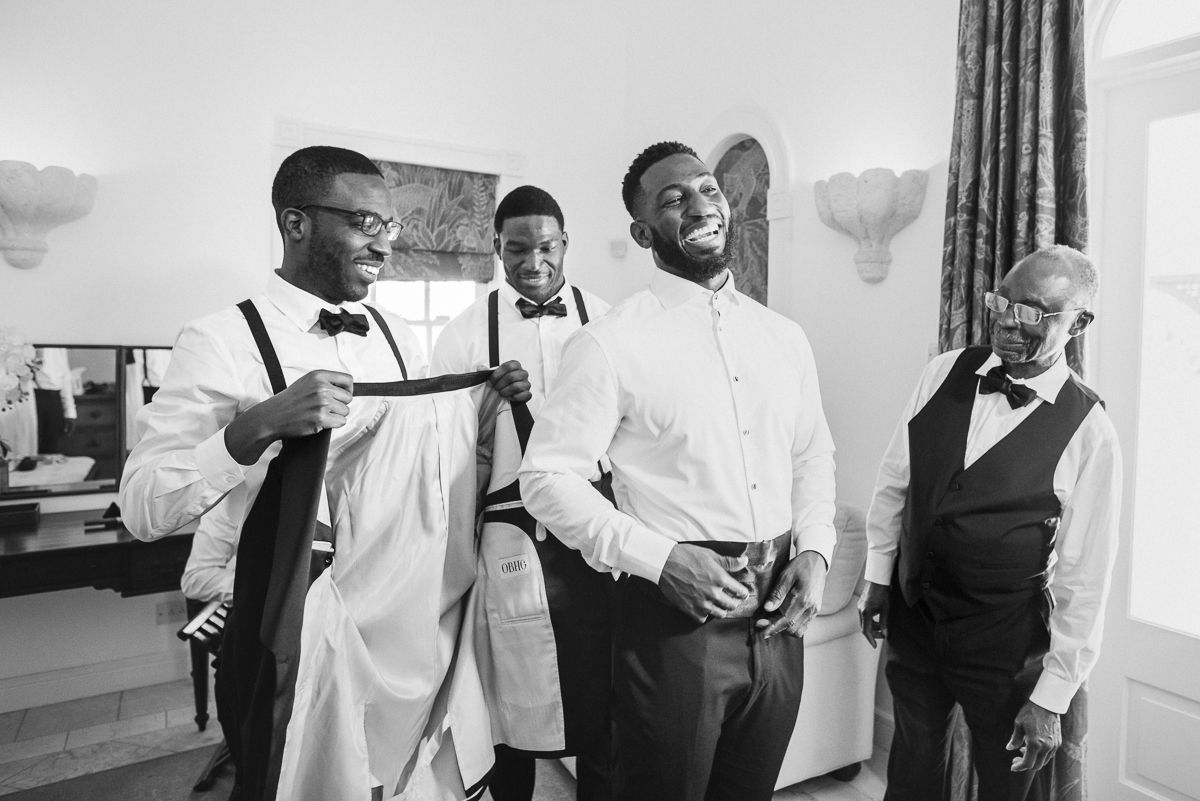 Groomsmen laughing and getting ready in black and white, adjusting suspenders before a wedding ceremony.