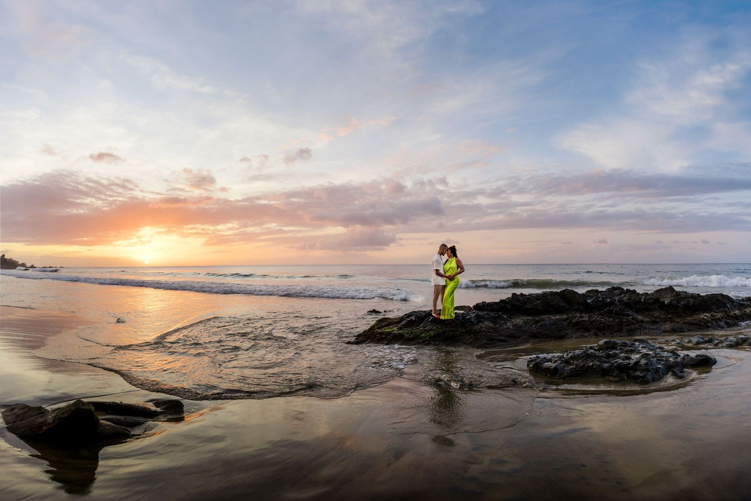 Person in yellow dress standing on rocks at a stunning beach sunset with colorful sky and ocean reflections.