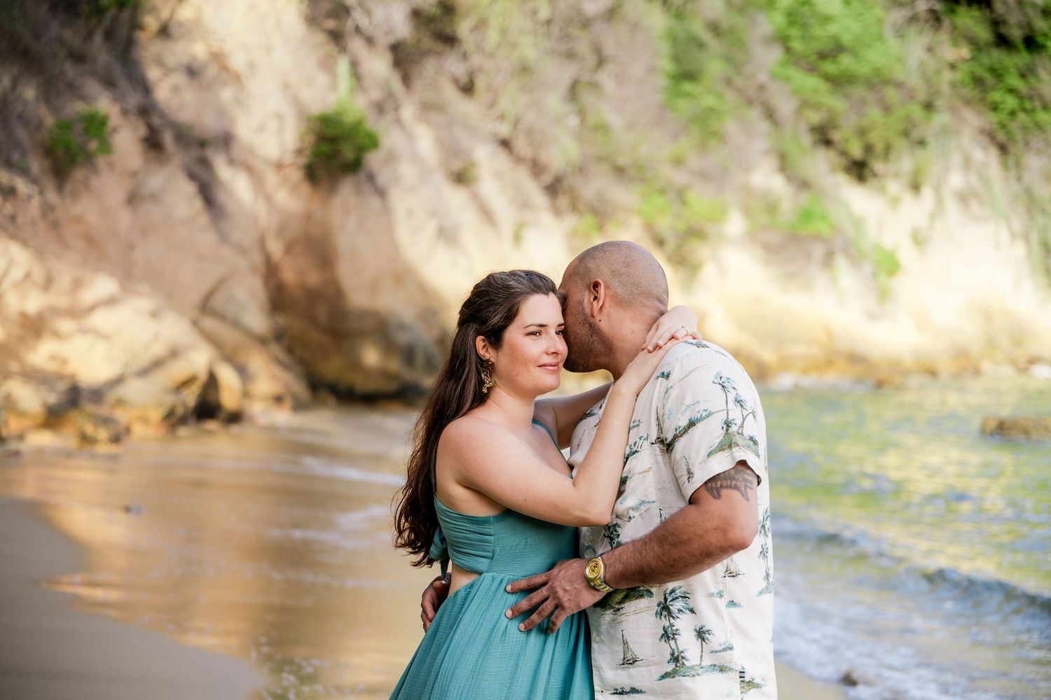 A couple embraces romantically on a scenic beach with rocky cliffs, woman wearing a teal dress.