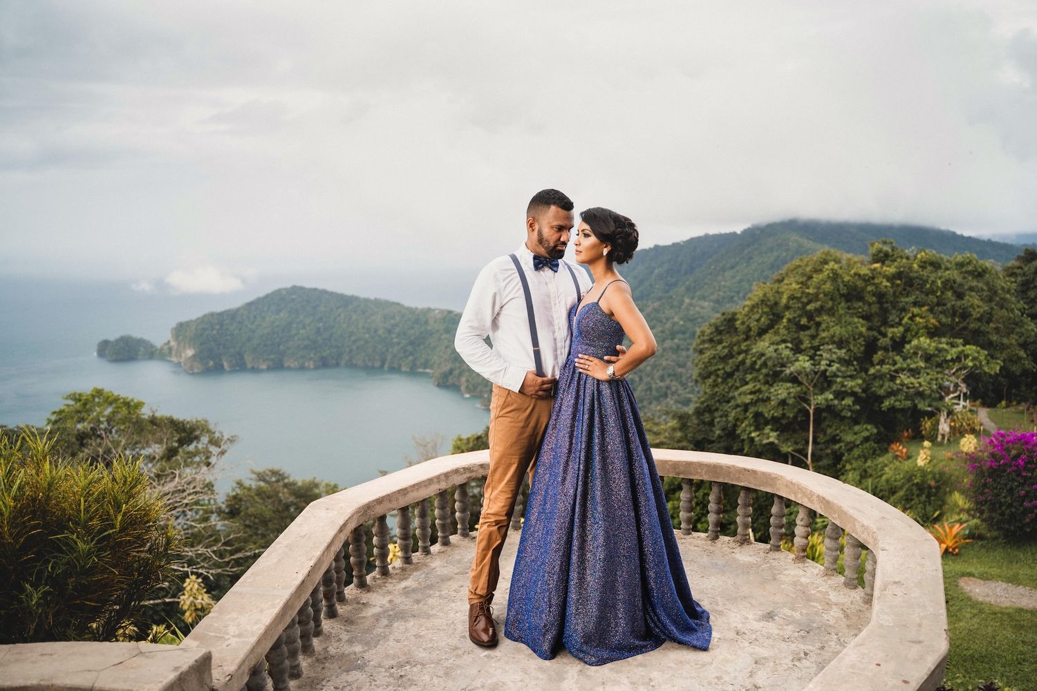 Couple in elegant attire poses on a stone balcony overlooking a lush tropical bay under cloudy skies.