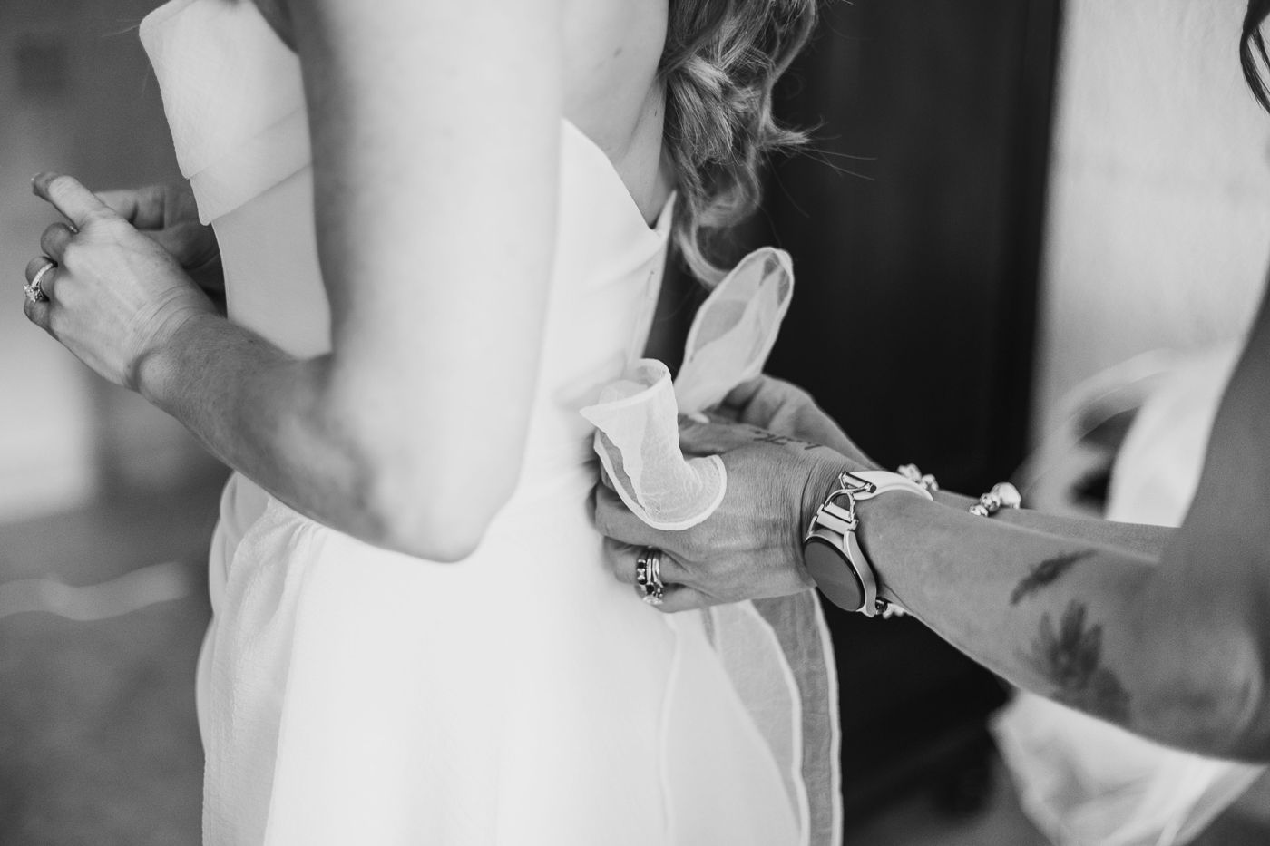 Black and white photo of a bride getting her wedding dress buttoned up by a helping hand.