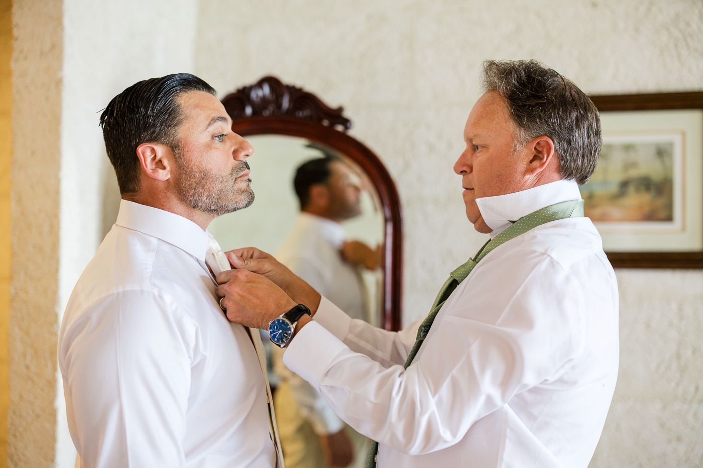 Two men in white dress shirts prepare for a wedding, one helping the other adjust his tie near an ornate mirror.