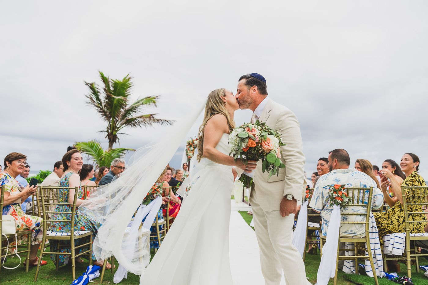 Bride and groom share first kiss at outdoor beach wedding ceremony with guests cheering and palm trees in background.