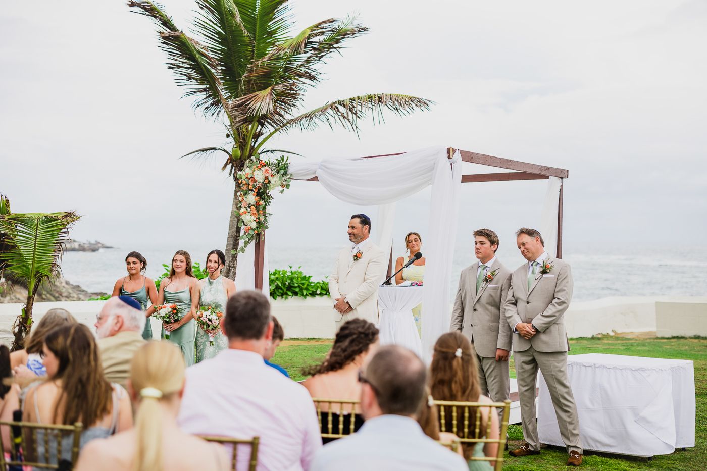 Outdoor beach wedding ceremony with bridal party under floral arch, palm trees, and ocean views on a cloudy day.