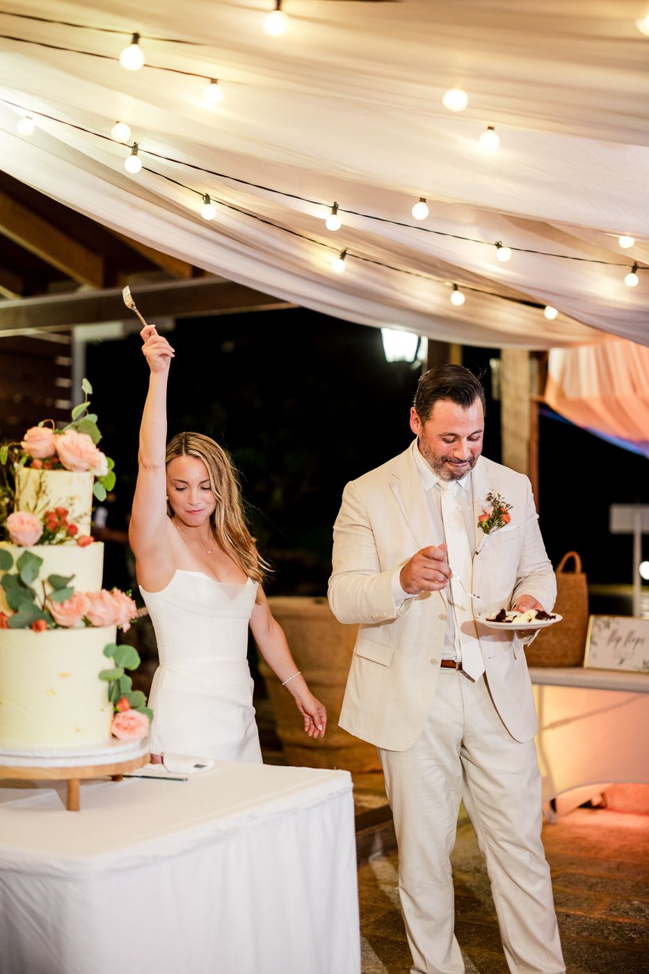 Bride and groom cutting their wedding cake under string lights and draped fabric at an outdoor evening reception.