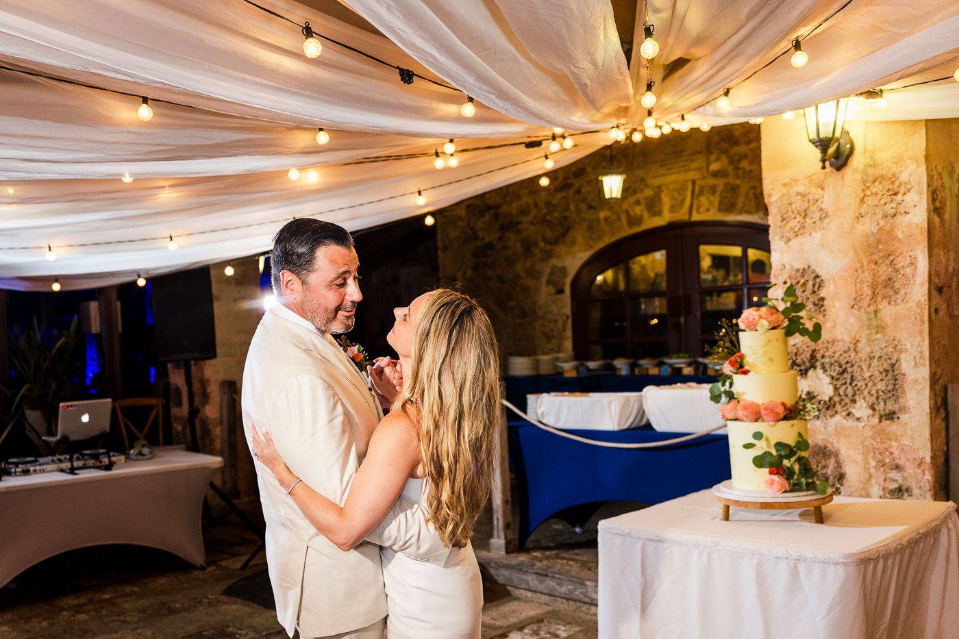 Bride and groom share a joyful moment cutting their wedding cake at an elegant outdoor reception with string lights.