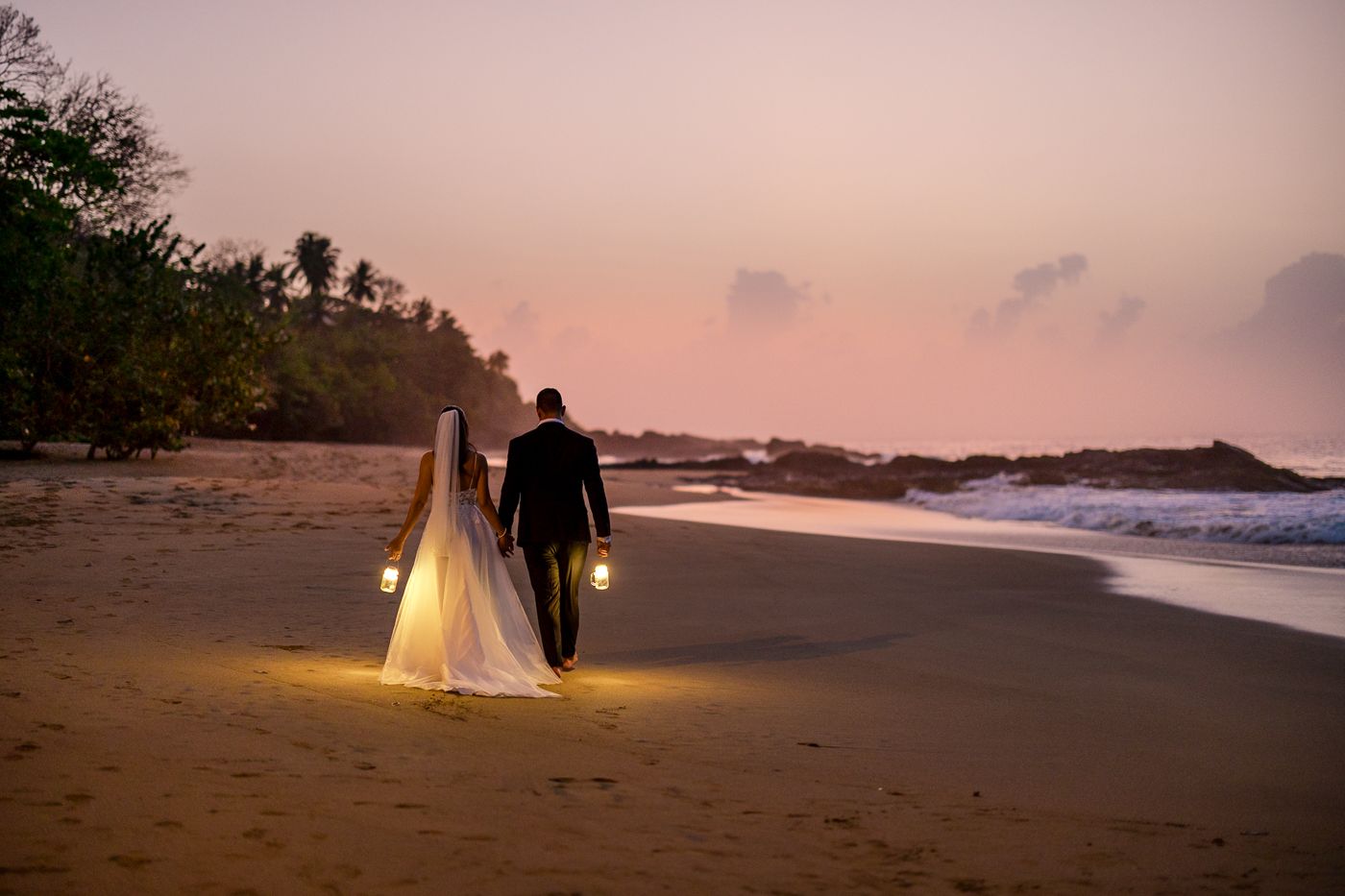 Bride and groom walk hand in hand along a beach at sunset, carrying glowing lanterns on their wedding day.