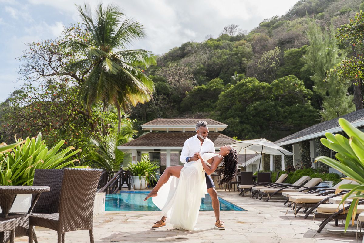 Couple dancing romantically by a tropical resort pool with palm trees and lush green hills in the background.