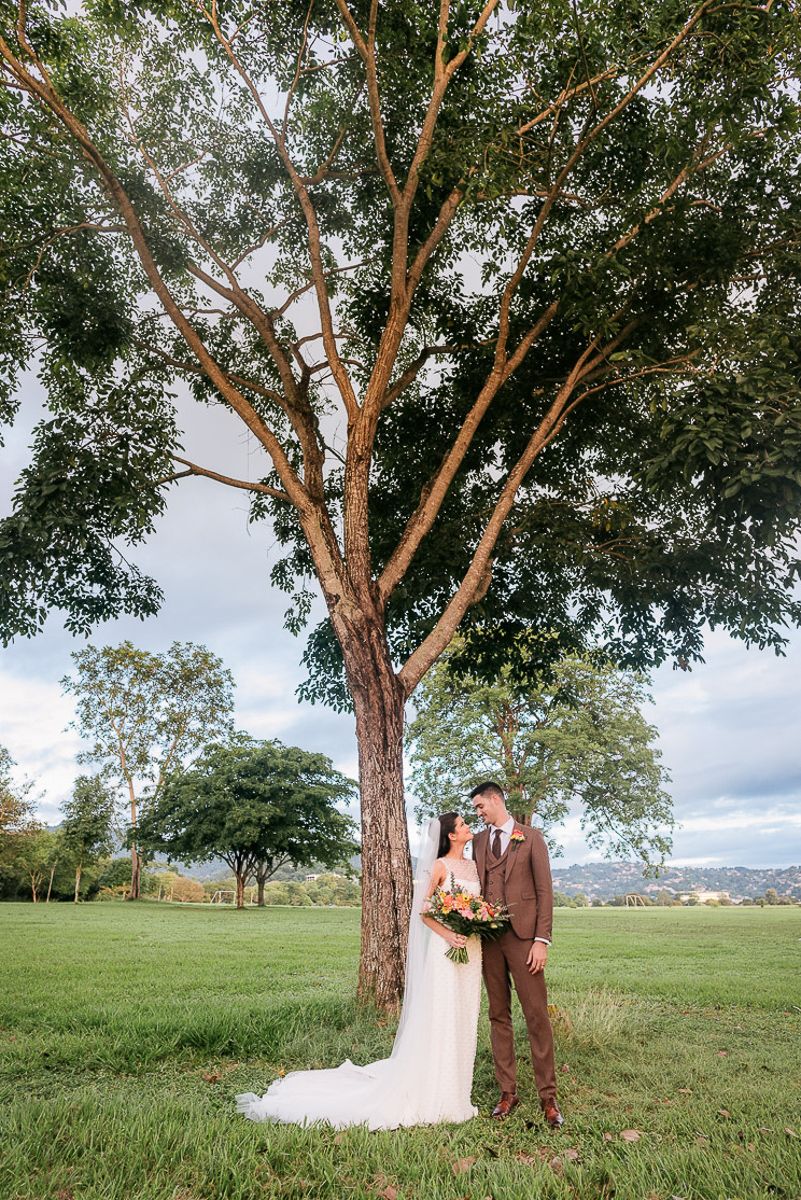 Bride and groom standing under a large tree in a green field on their wedding day.