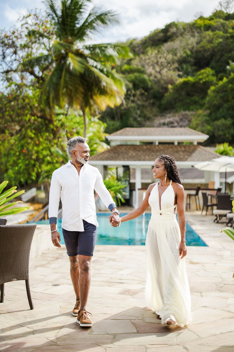 Couple holding hands walking by tropical pool, wearing white outfits, with palm trees and villa in background.