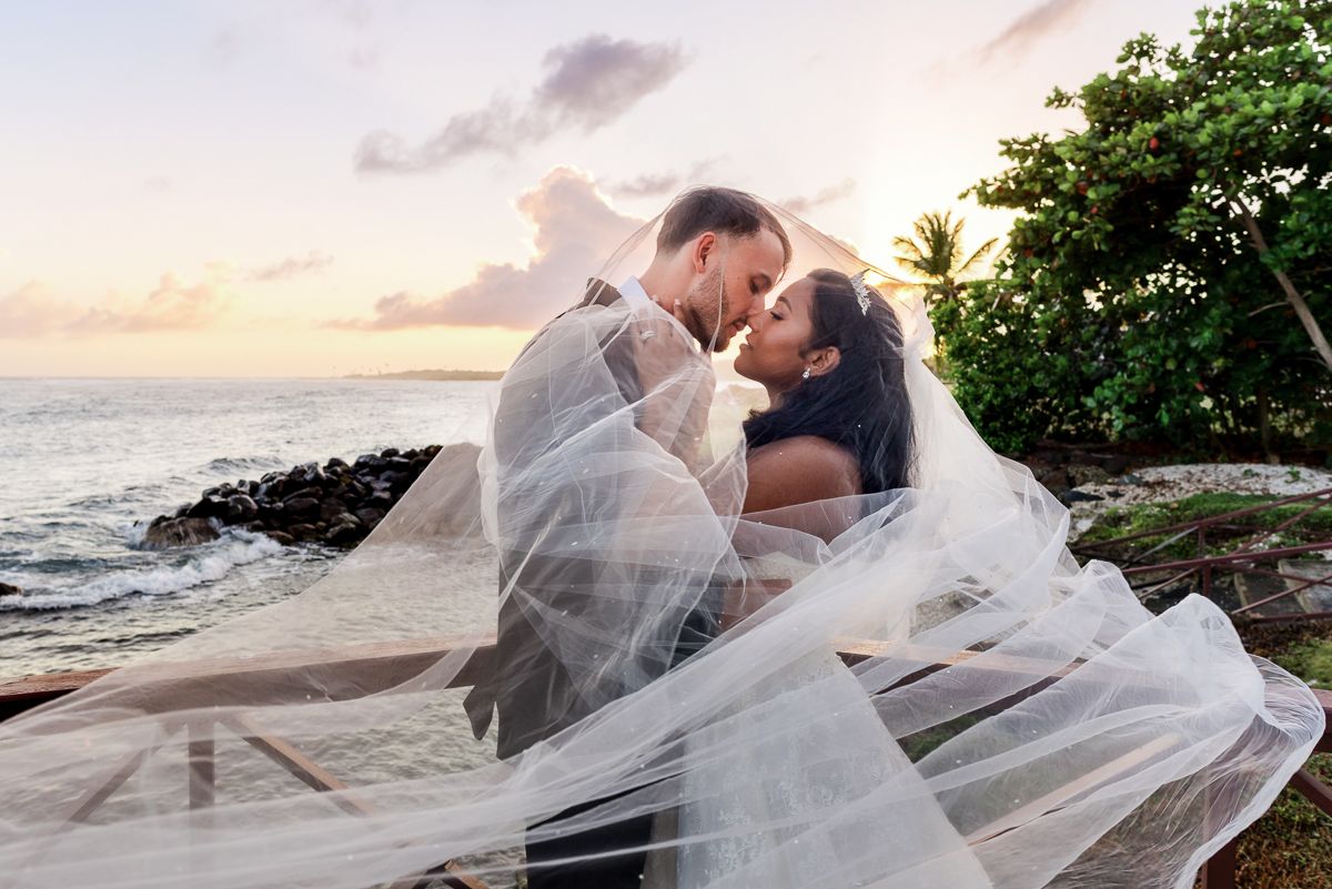 Newlywed couple sharing a romantic sunset kiss on a tropical beach, bride's veil flowing in the ocean breeze.