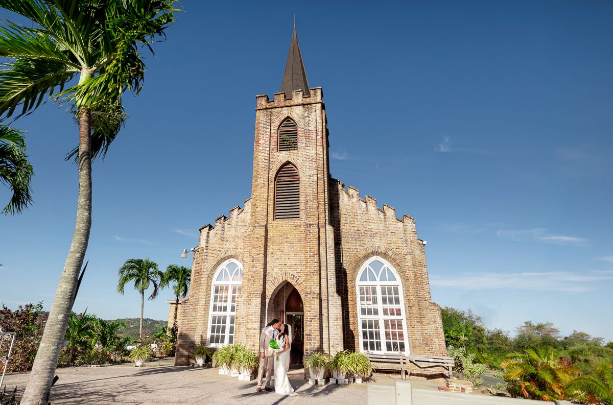 Couple posing in front of a historic stone church with arched windows and bell tower surrounded by palm trees.