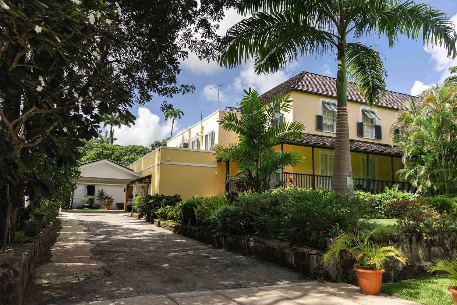 A curved driveway leads through a lush tropical garden with palm trees and yellow stucco buildings in the background.